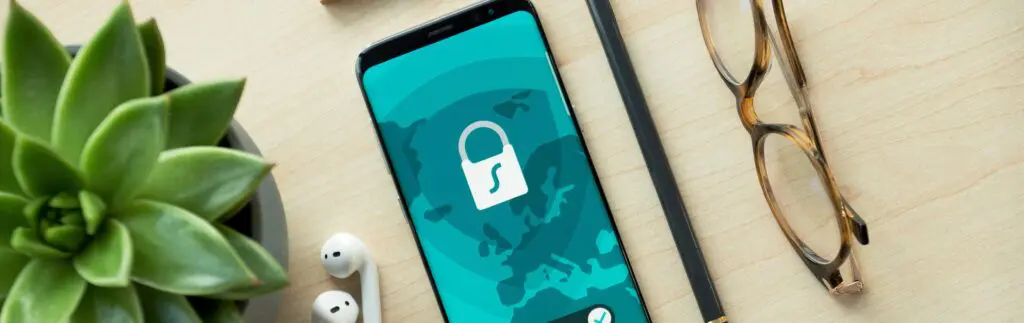 In der Mitte des Bildes ist ein Mobiltelefon mit einem Bild von einem Schloss darauf. Rund um das Mobiltelefon sind Kopfhörer, ein Stift, eine Brille und eine Pflanze.