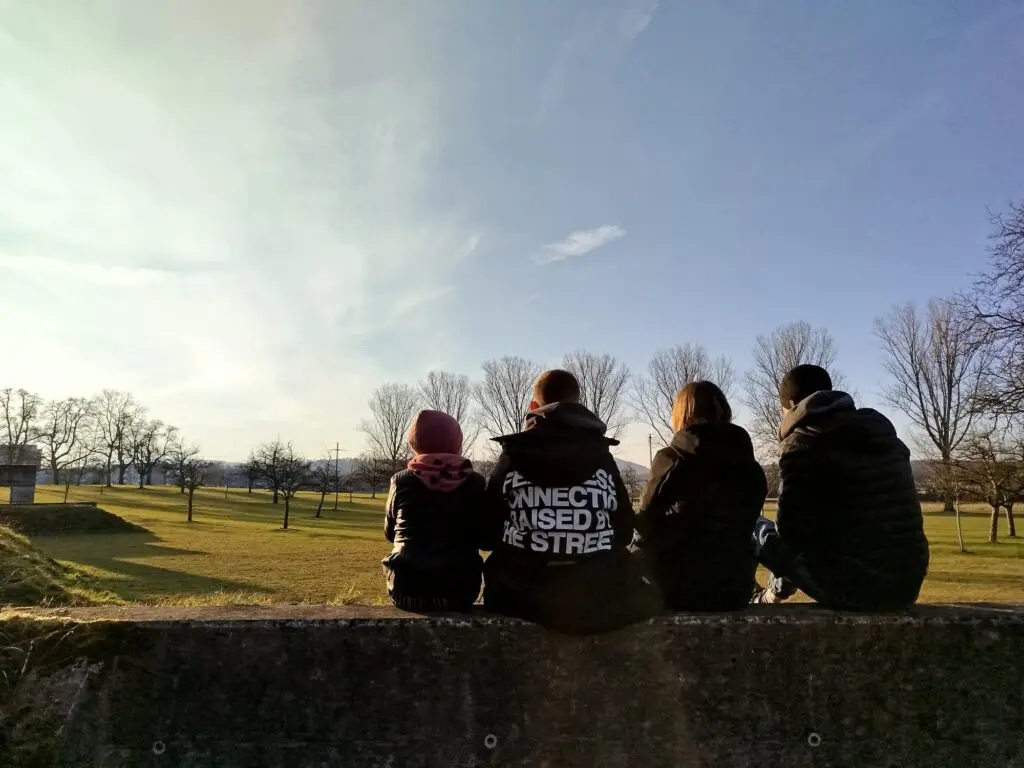 Zwei Kinder, eine Frau und ein Mann sitzen zusammen auf einer Mauer. Man sieht sie von hinten.