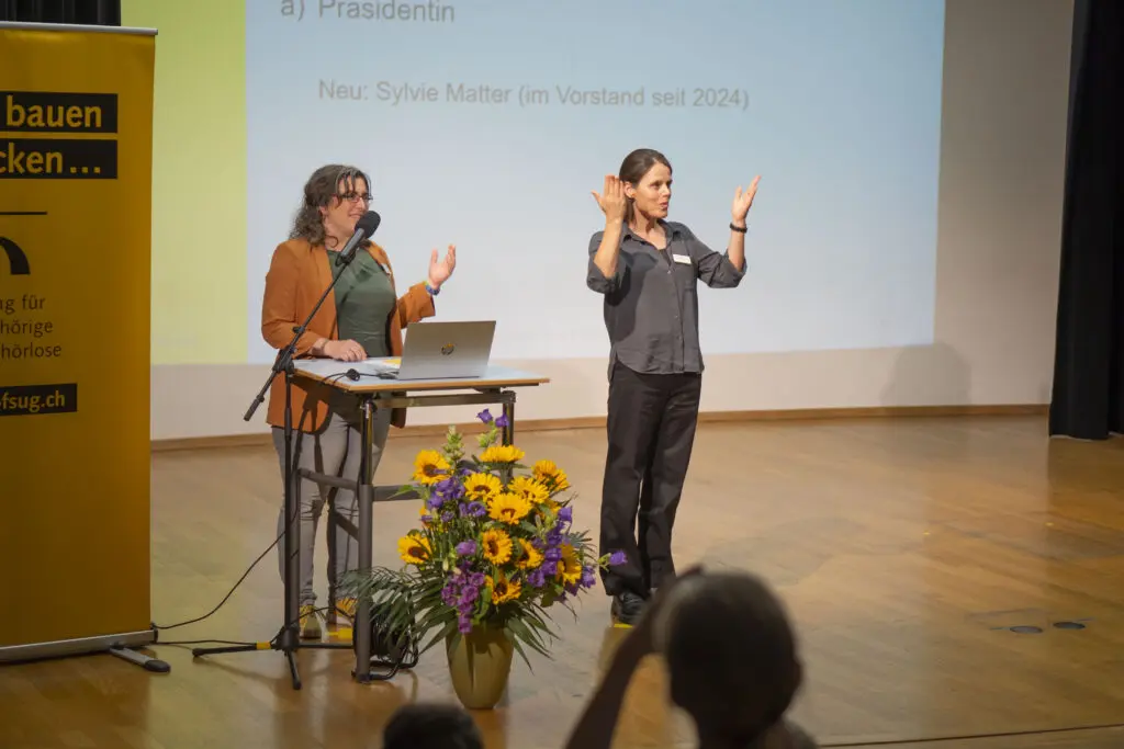 Eine Frau mit dunklen Haaren steht hinter einem Podium auf einer Bühne und spricht ins Mikrofon. Rechts von ihr ist eine Gebärdensprachdolmetscherin zu sehen, die gebärdet.