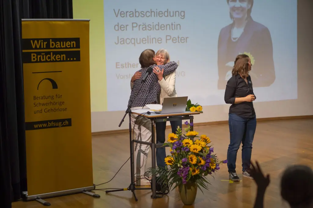 Eine blonde und eine braunhaarige Frau stehen auf einer Bünde hinter einem Podium und umarmen sich. Daneben steht eine Dolmetscherin, die von der Kamera wegschaut.