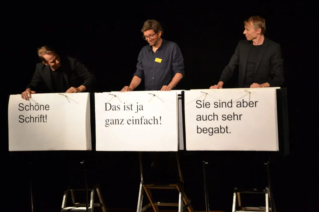 Es stehen drei Männer, die Plakate vor ihnen haben, auf einer Bühne. Auf dem Plakat links steht "Schöne Schrift!", auf dem mittleren "Das ist ja ganz einfach!" und auf dem rechten ""Sie sind aber auch sehr begabt".