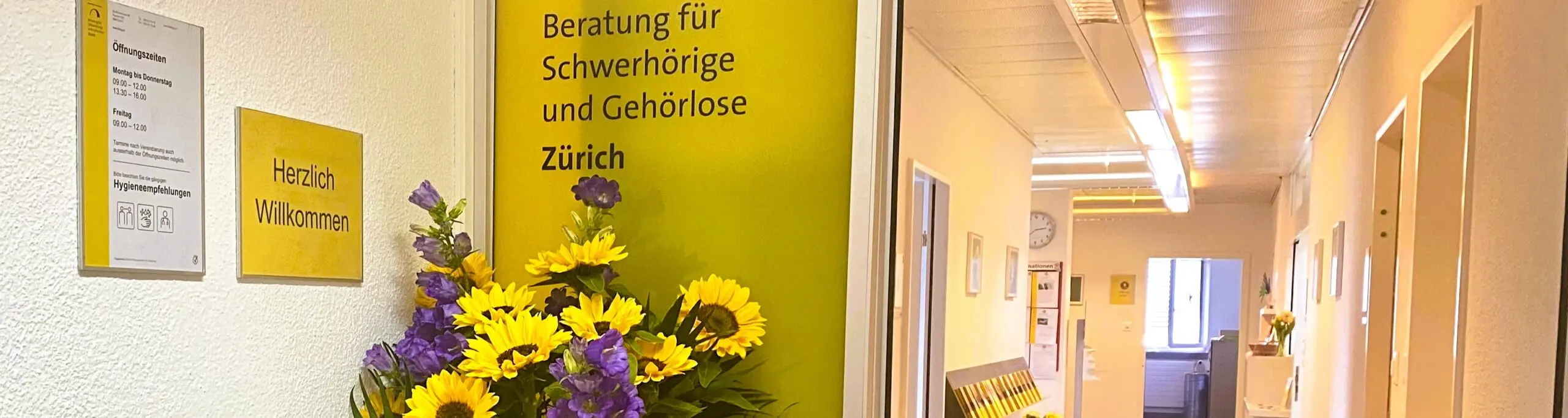 Es ist eine gelbe offene Schiebetür zu sehen. Davor ist ein Blumenstrauss. Durch die offene Tür sieht man einen Gang.
