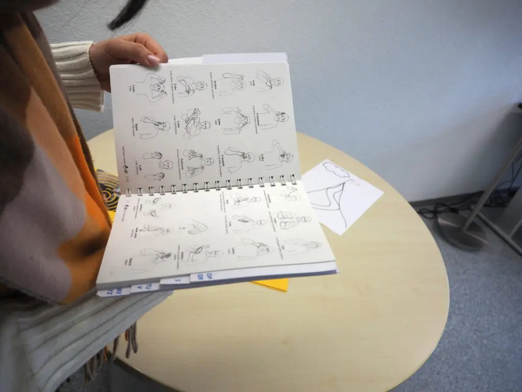 Eine Person hält ein offenes Buch in der Hand. Das Buch zeigt Piktogramme von Gebärden mit den passenden Begriffen.