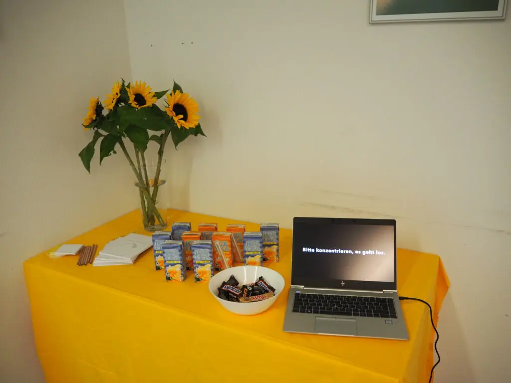 Auf einem Tisch mit gelbem Tischtuch stehen eine Blume, Eistee-Päckchen, Schokolade, Stifte, Papier, Post-Its und ein Laptop auf dessen Bildschirm "Bitte konzentrieren, es geht los" steht.