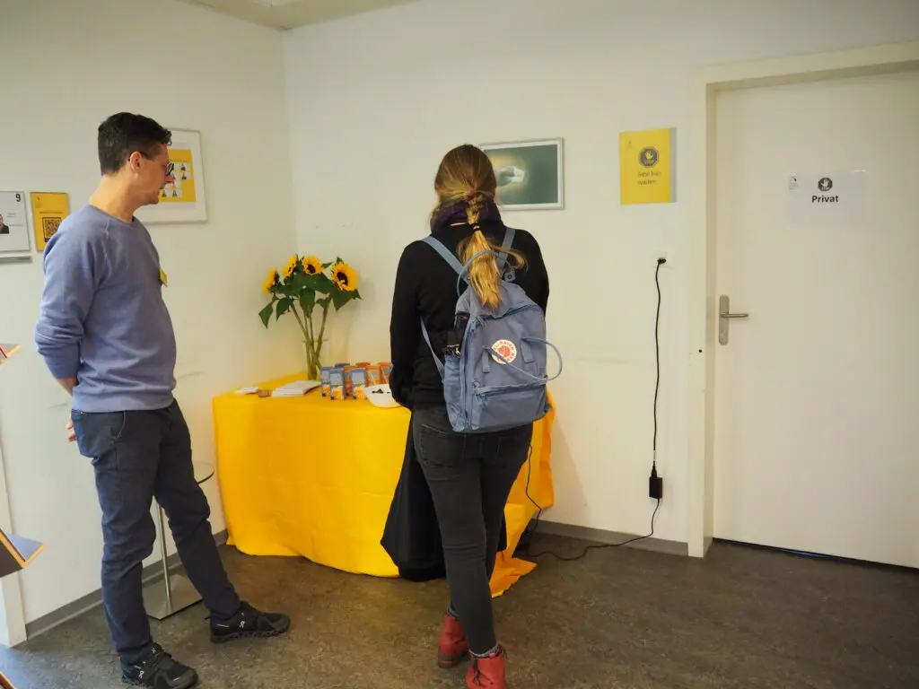 Eine Frau steht mit dem Rücken zur Kamera und neben ihr steht ein Mann, der zu ihr gerichtet steht. Beide schauen auf einen Tisch mit gelbem Tischtuch. An der Wand hinter dem Tisch sind verschiedene Bilder.