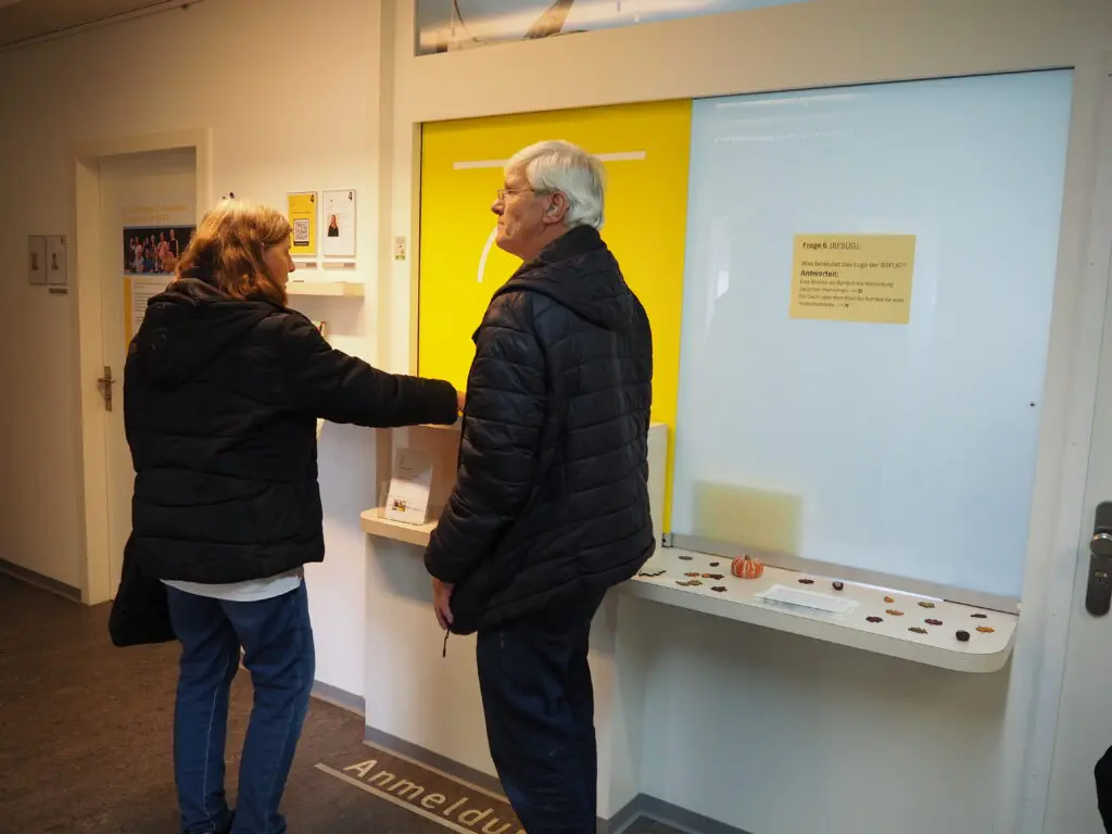 Eine Frau steht mit dem Rücken zur Kamera und ein Mann steht seitlich zur Kamera. Der Mann schaut geradeaus und die Frau schaut auf den Tresen des Empfangs.