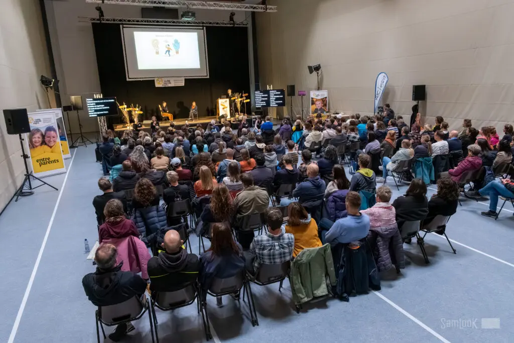 Auf dem Bild sind Zuschauer von hinten zu sehen. Sie schauen auf eine Bühne, auf der zwei Personen sitzen. Oberhalb der Bühne ist ein Bildschirm mit Piktogrammen von einer Glühbirne, einer Person mit Schirm und einem Handschuh.