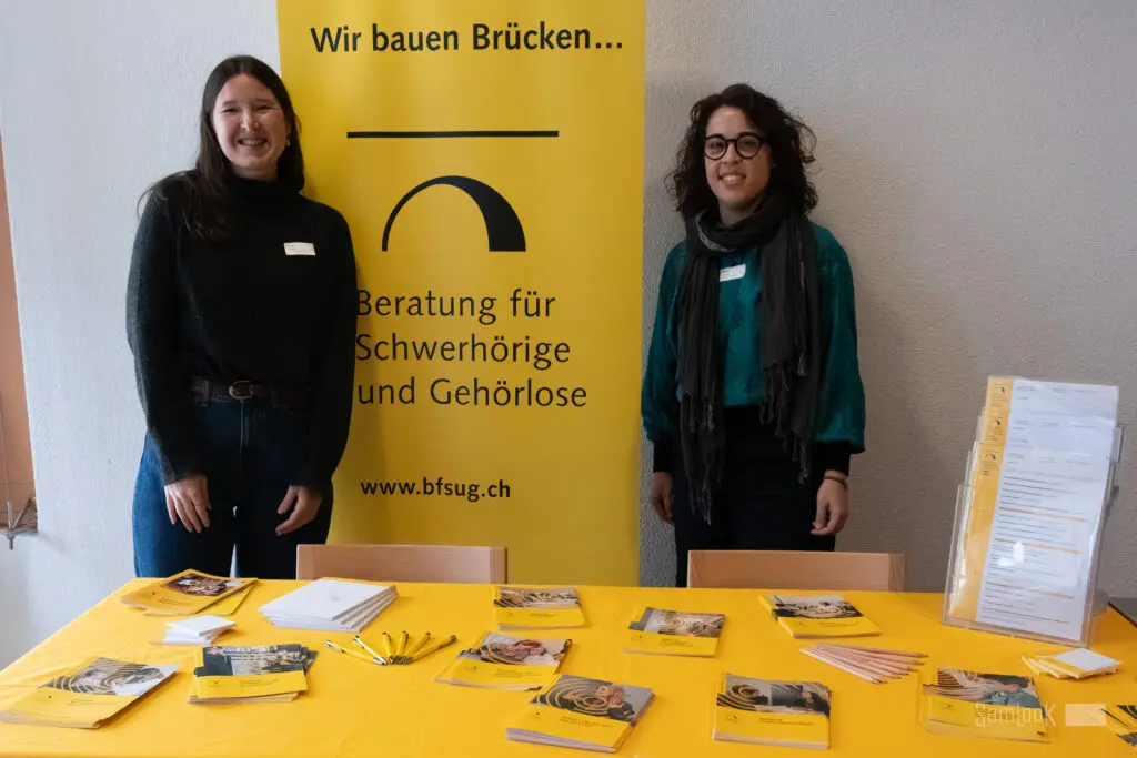 Zwei Frauen stehen links und rechts von einem gelben Banner auf dem "Wir bauen Brücken... Beratung für Schwerhörige und Gehörlose" steht. Vor ihnen steht ein Tisch mit gelbem Tischtuch, auf dem Stifte, Post-Its, Notizblöcke, Informationsblätter und verschiedene Broschüren sind.