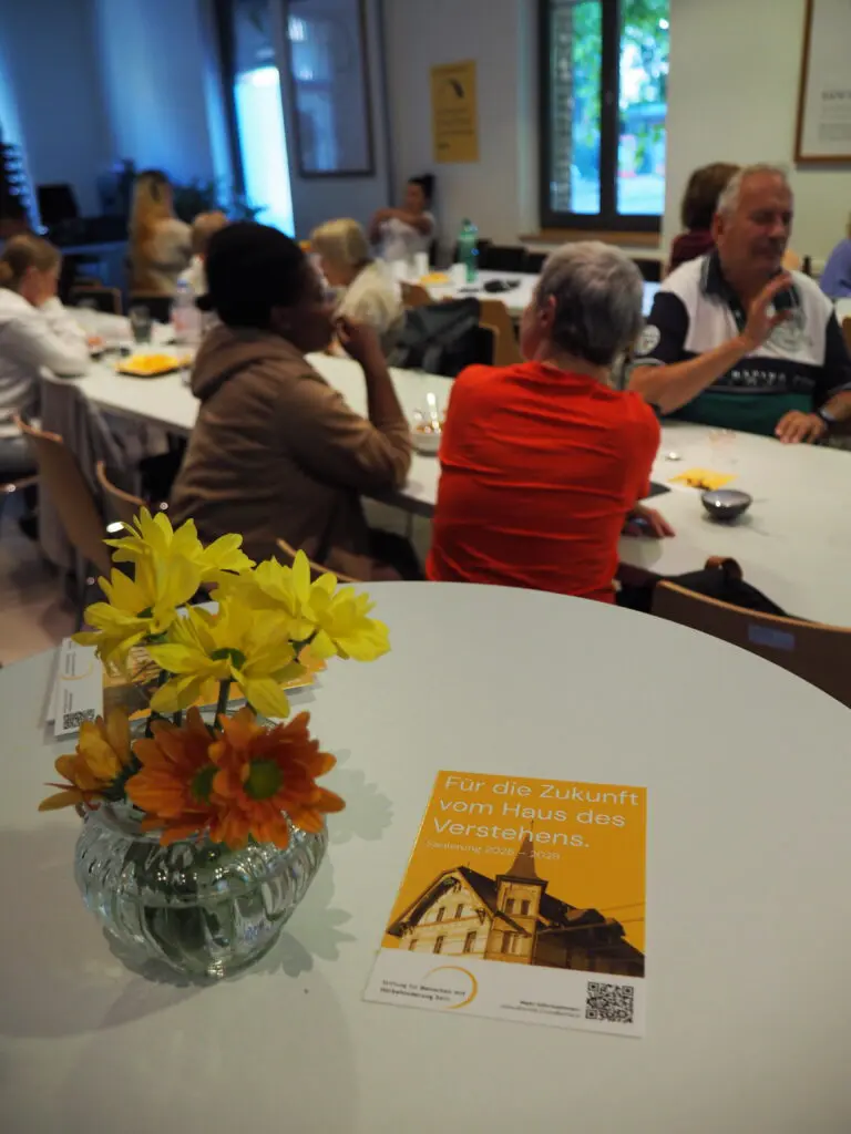 Open House & Sommerfest BFSUG Bern Tisch mit Flyer: Für die Zukunft vom Haus des Verstehens und hinter dem Tisch sieht man mehrere Personen an einem Tisch die sich austauschen.