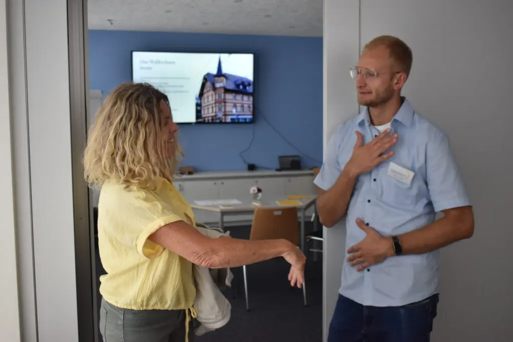 Open House & Sommerfest BFSUG Bern. Eine Frau und ein Mann unterhalten sich in Gebärdensprache vor einem Bildschirm mit einem Bild vom Walkerhaus.