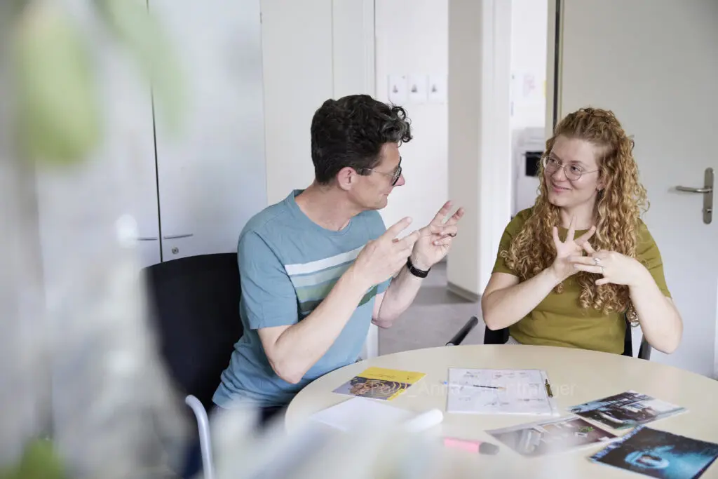 Eine Frau und ein Mann sitzen zusammen an einem runden Tisch und gebärden. Auf dem Tisch liegen mehrere Dokumente und Broschüren.