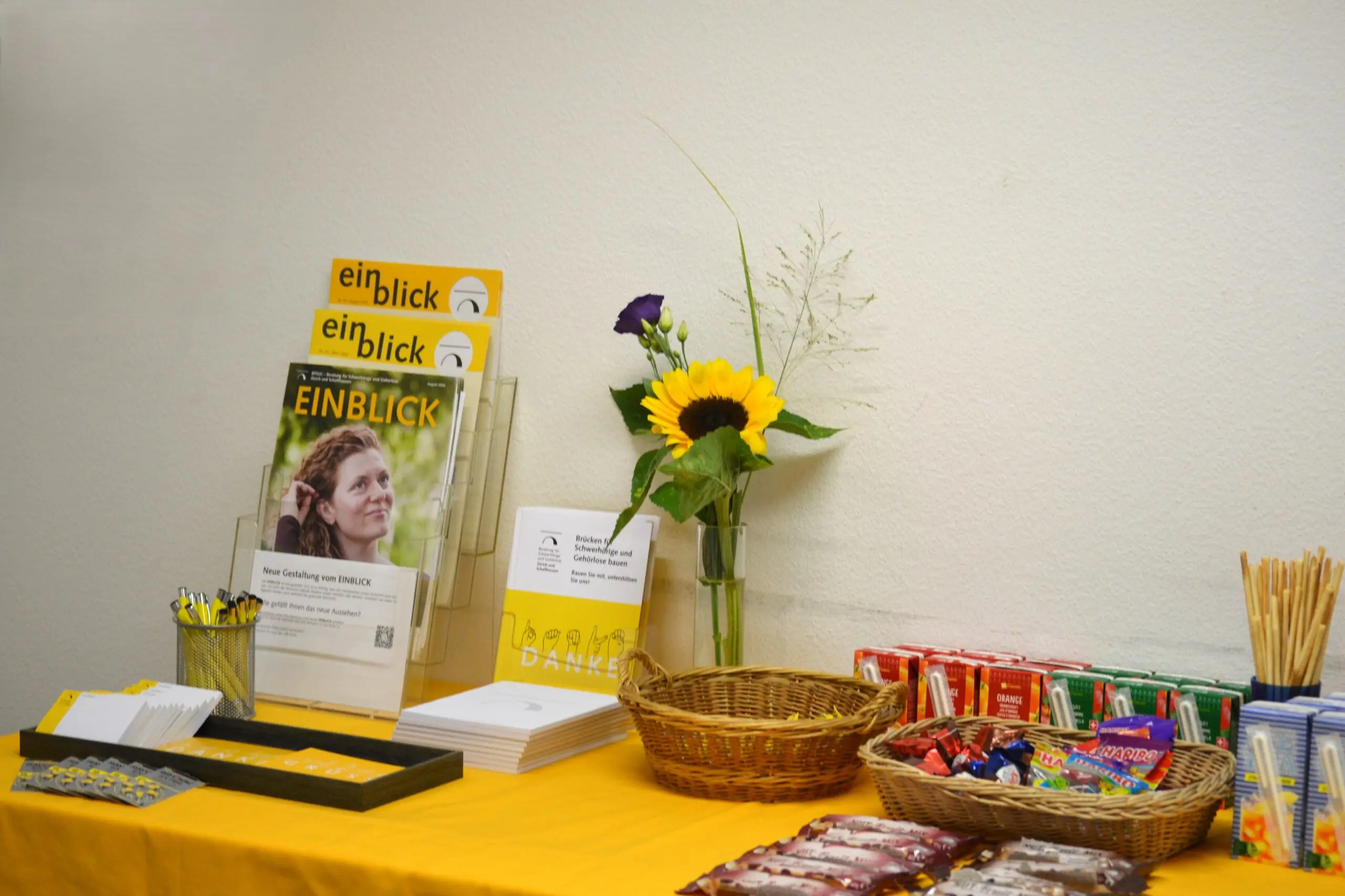 Auf einem Tisch mit gelbem Tischtuch stehen Post-Its, Kugelschreiber, Broschüren, Sonnenblumen und Snacks.