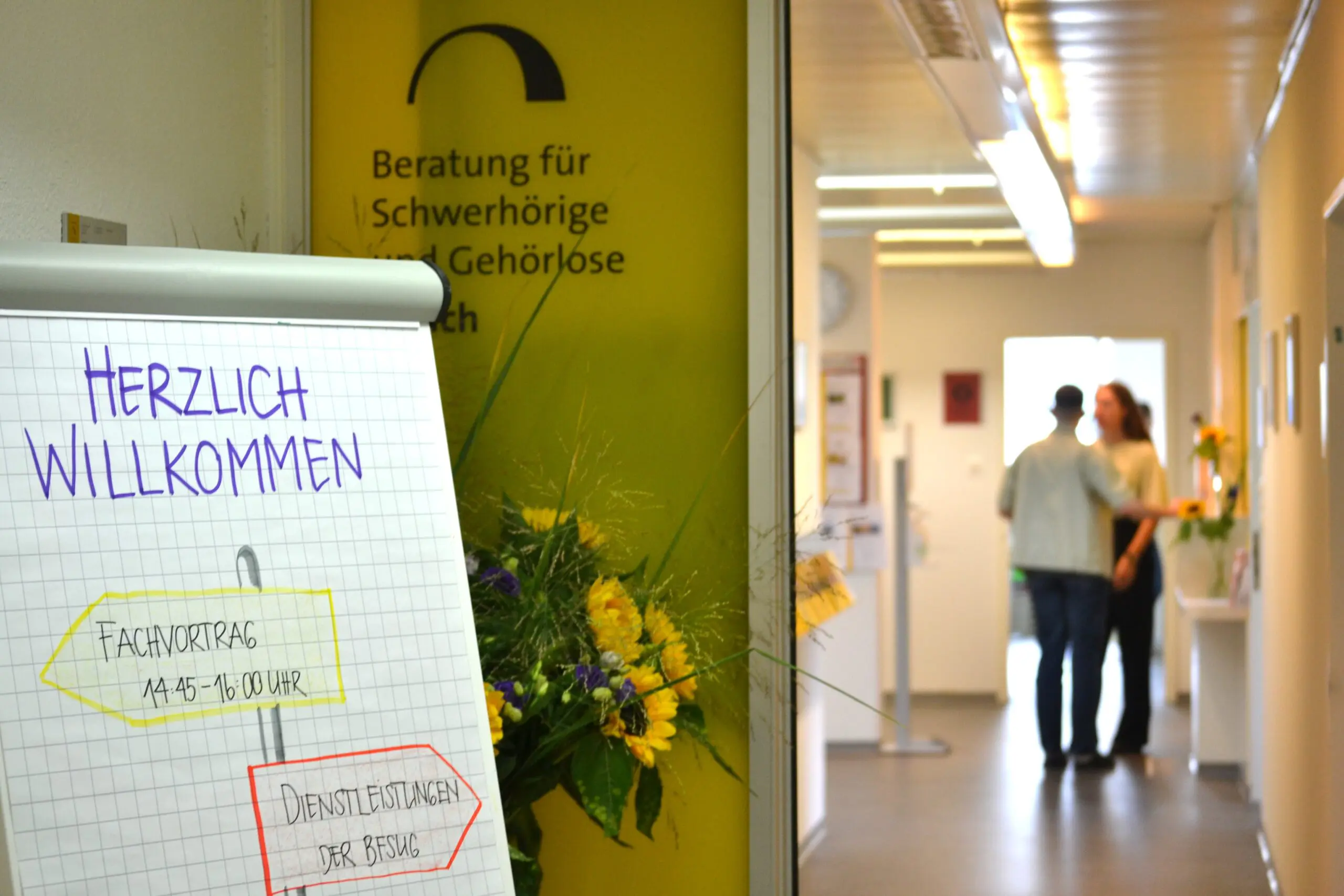 Es ist eine offene, gelbe Schiebetür mit der Aufschrift "Beratung für Schwerhörige und Gehörlose Zürich" zu sehen. Davor steht ein Schild mit der Aufschrift "Herzlich Willkommen. Fachvortrag. Dienstleistungen der BFSUG". Hinter der Schiebetüre sind zwei Personen, die miteinander reden. Die Personen sind unscharf.
