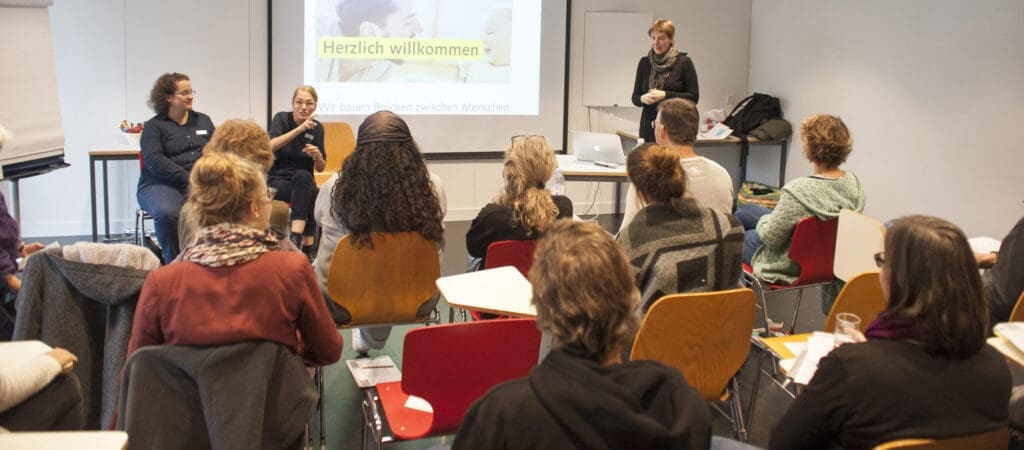 Eine Frau hält einen Vortrag und zwei Gebärdensprachdolmetscherinnen übersetzen. Vom Publikum, das auf Stühlen sitzt, ist der Rücken ersichtlich.
