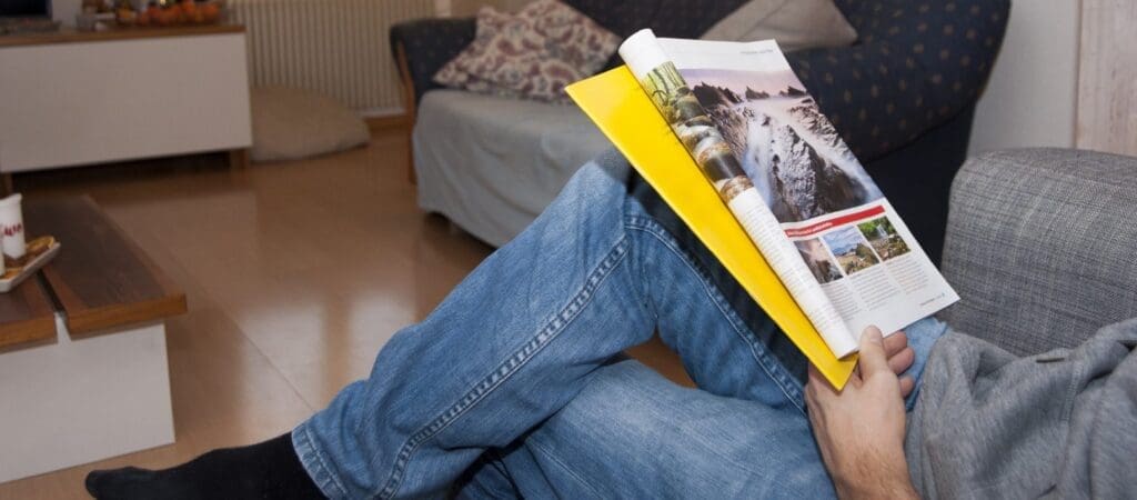 Ein Mann sitzt mit überkreuzten Beinen auf einem Sofa und liest eine Zeitschrift, in der ein Bild von einem Berg zu sehen ist.
