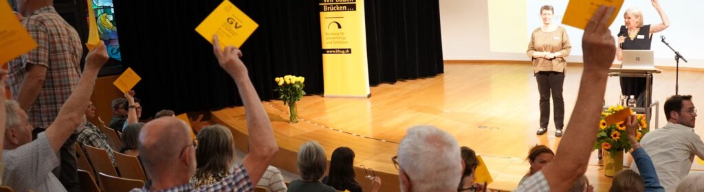 Abstimmung an der Generalversammlung 2023, diverse Personen heben die gelbe Stimmkarte, auf der Bühne referiert die Präsidentin und die Gebärdensprach Dolmetscherin übersetzt.