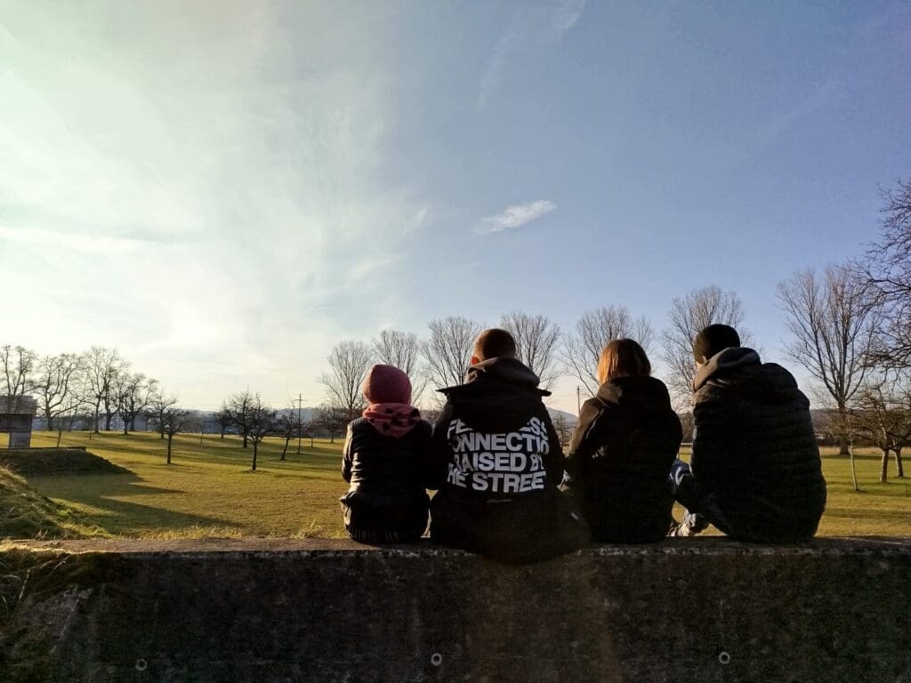 Zwei Kinder, eine Frau und ein Mann sitzen zusammen auf einer Mauer. Man sieht sie von hinten.