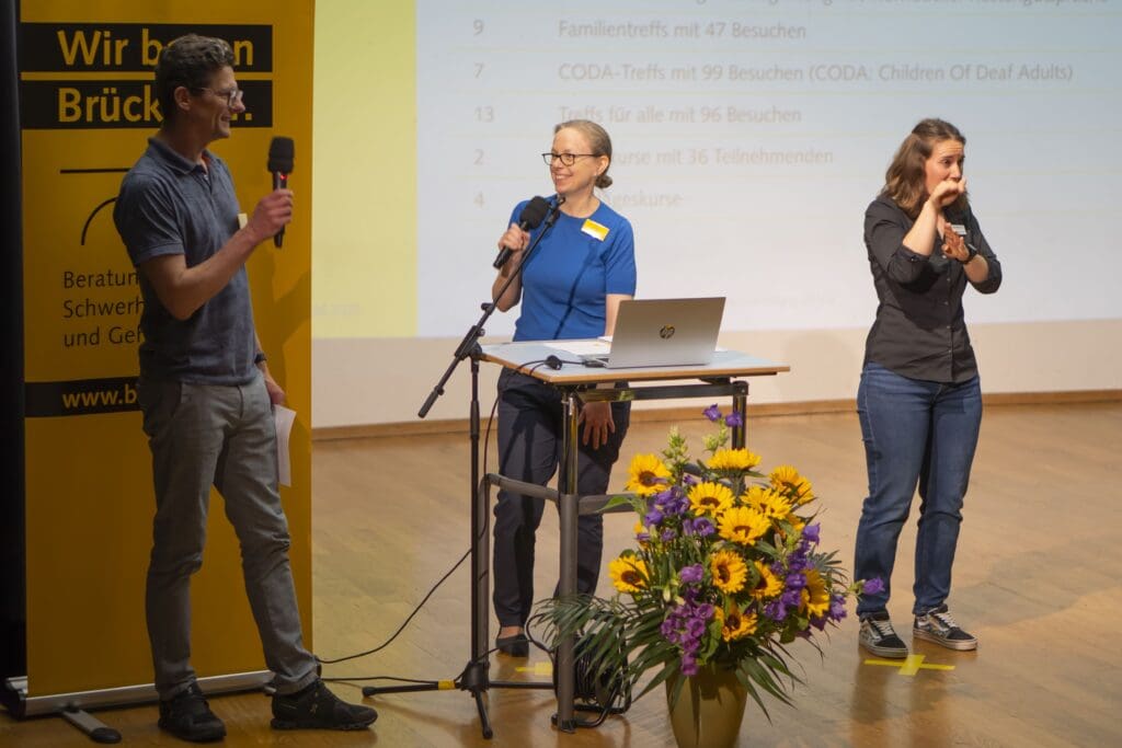 Ein Mann und eine Frau stehen auf einer Bühne, schauen sich gegenseitig an und sprechen in ein Mikrofon. Rechts von ihnen steht eine Gebärdensprachdolmetscherin, die übersetzt.