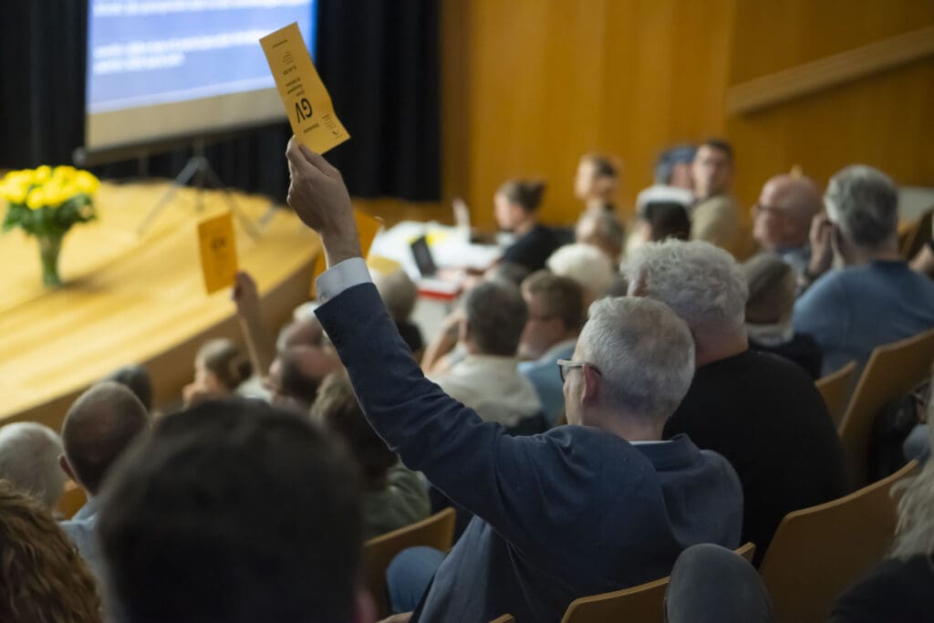 Es ist ein Publikum zu sehen. Einige Leute halten ein gelbes Papier in die Luft, auf dem "GV" steht.