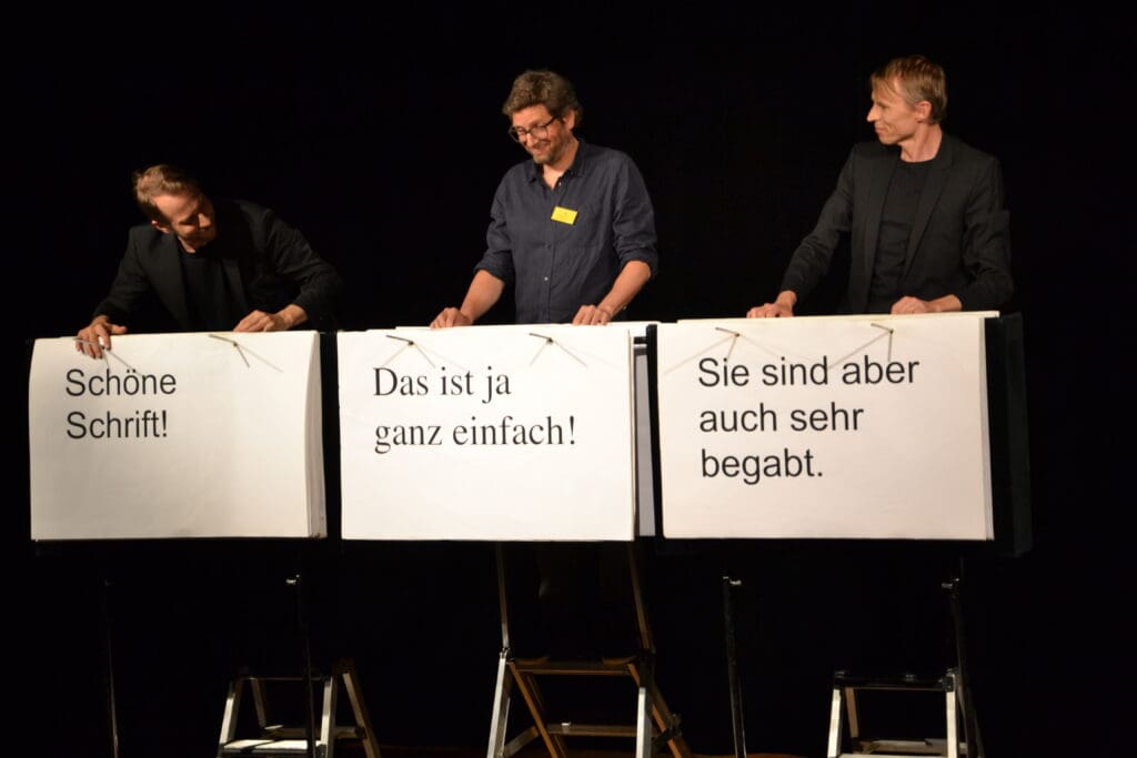 Es stehen drei Männer, die Plakate vor ihnen haben, auf einer Bühne. Auf dem Plakat links steht "Schöne Schrift!", auf dem mittleren "Das ist ja ganz einfach!" und auf dem rechten ""Sie sind aber auch sehr begabt".