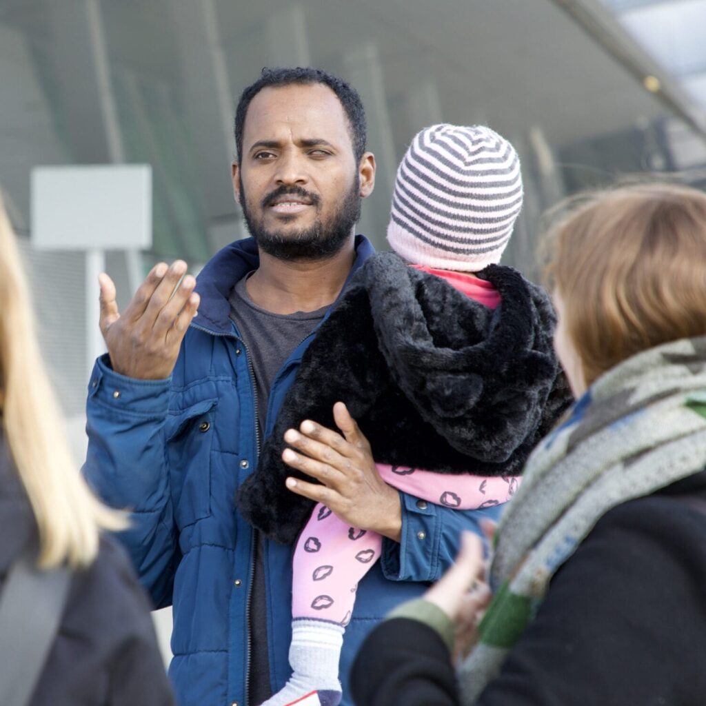 Ein Mann hält ein Kind im Arm. Er unterhält sich in Gebärdensprache mit zwei Frauen, die man von hinten sieht.