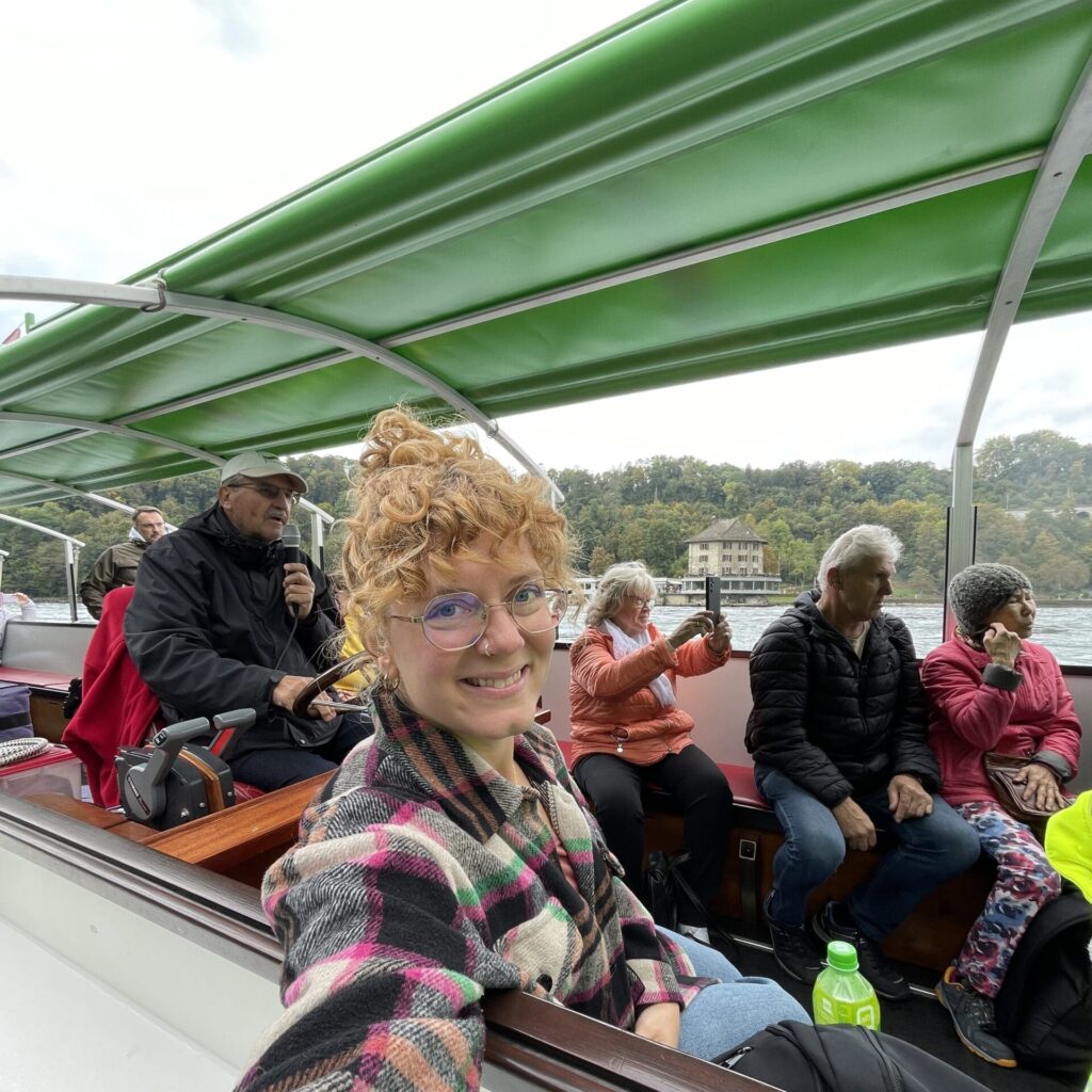 Eine Frau macht auf einem Boot ein Selfie und lächelt in die Kamera. Im Hintergrund sitzen weitere Personen im Boot.