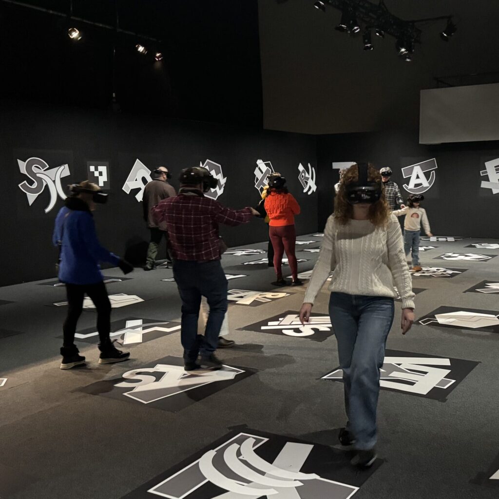 Mehrere Menschen gehen durch einen dunklen Raum, auf dessen Boden verschiedene Zeichen sind. Alle Personen tragen VR-Brillen.