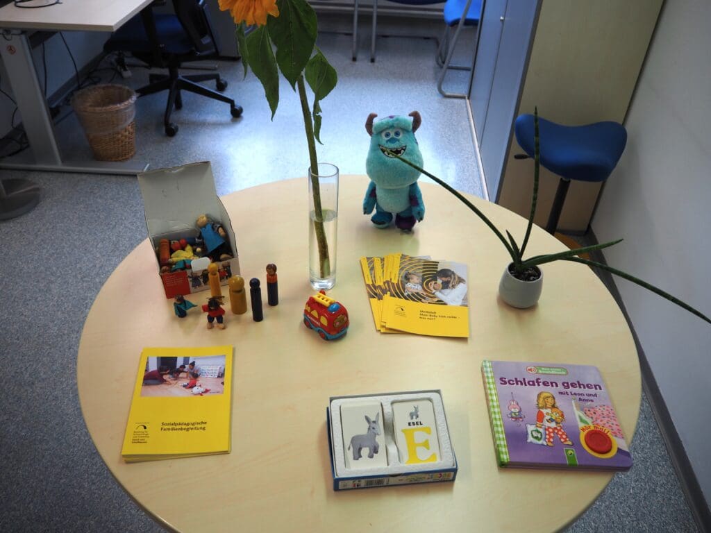 Auf einem runden Tisch stehen eine Blume, Kinderspielzeug, Flyer mit den Aufschriften "Sozialpädagogische Familienbegleitung" und "Merkblatt mein Baby hört nichts - was tun?", ein Kinderbuch, ein Memory und eine Tischpflanze.